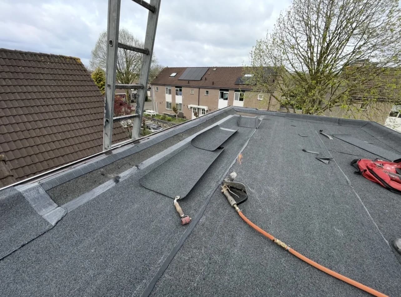Bitumen wordt gebrand langs de dakrand naast een pannendak, met ladder en brander